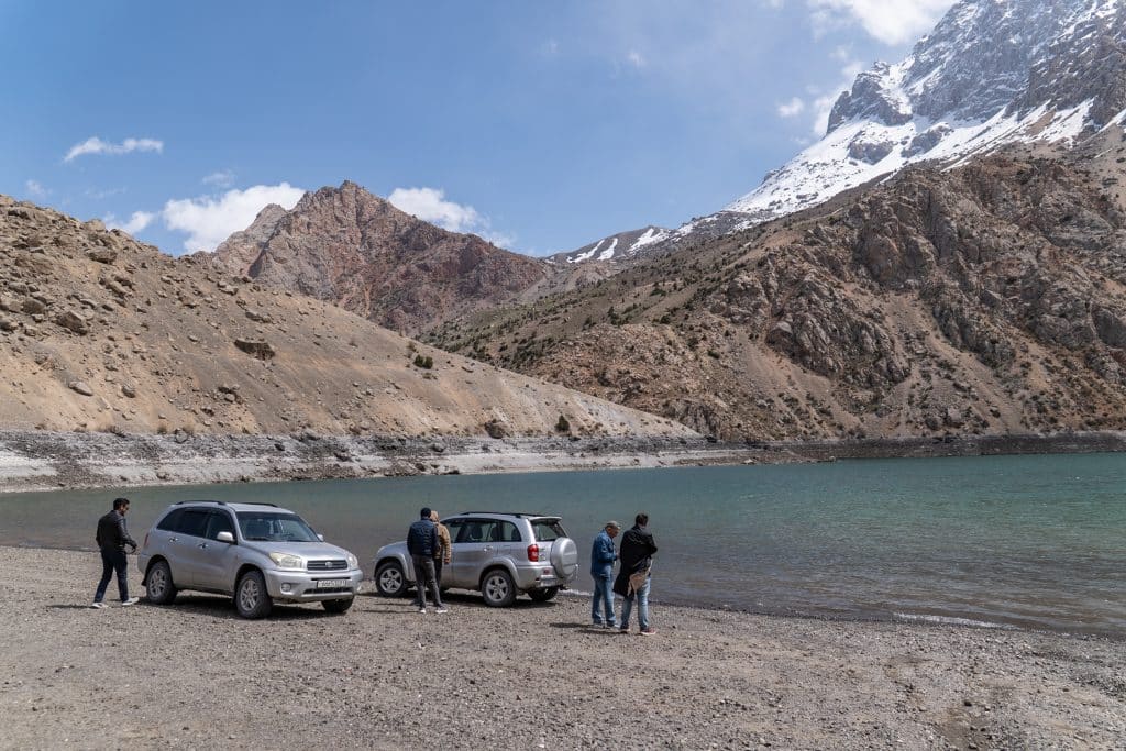 A group of people standing by two cars parked by a mountain lake in Tajikistan.