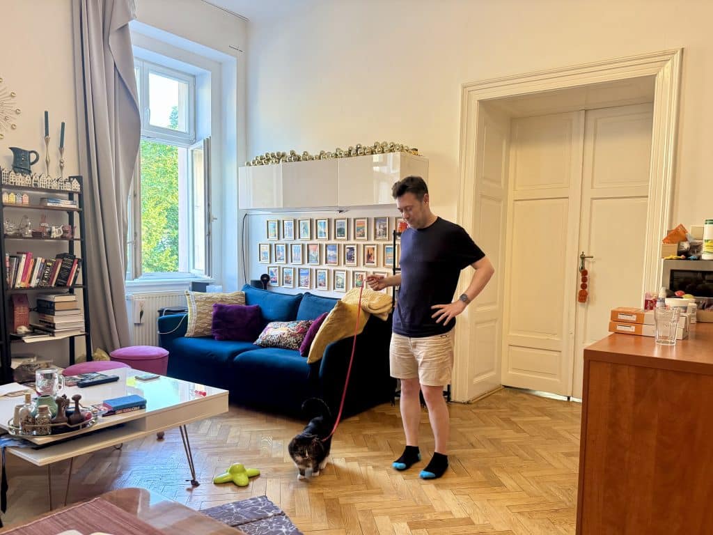 Charlie stands in his and Kate's living room, with the windows wide open. He holds a red leash attached to Lewis the little gray cat, who looks a bit disgruntled.