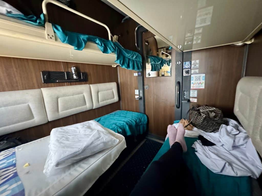 A sleeper train car. Kate's legs are stretched out on her bunk, and there is an upper bunk above each bed.