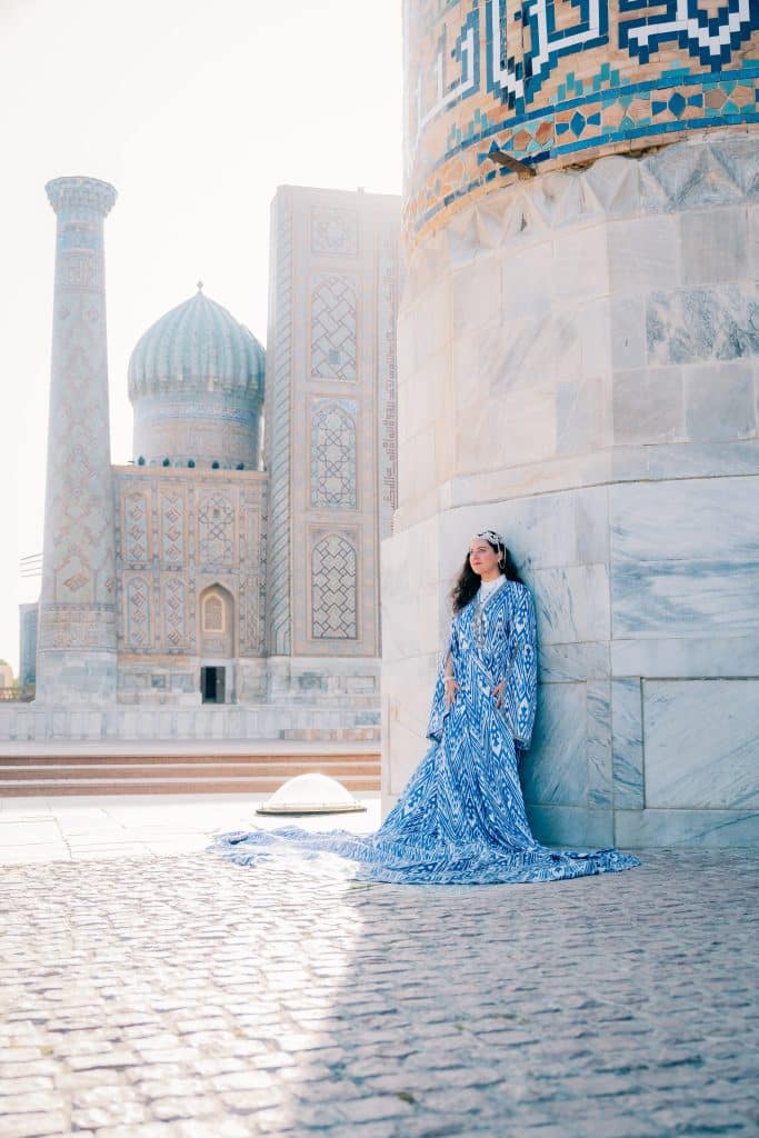 Kate in a long flowy blue Uzbek dress at the Registan, turquoise columns behind her.