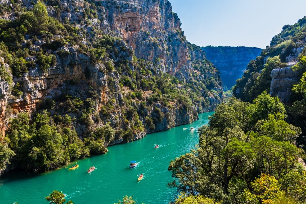 A gorge in Fance with bright green vegetation-covered walls, and a bright turquoise river snaking through it. The river is filled with people in kayaks and small boats.
