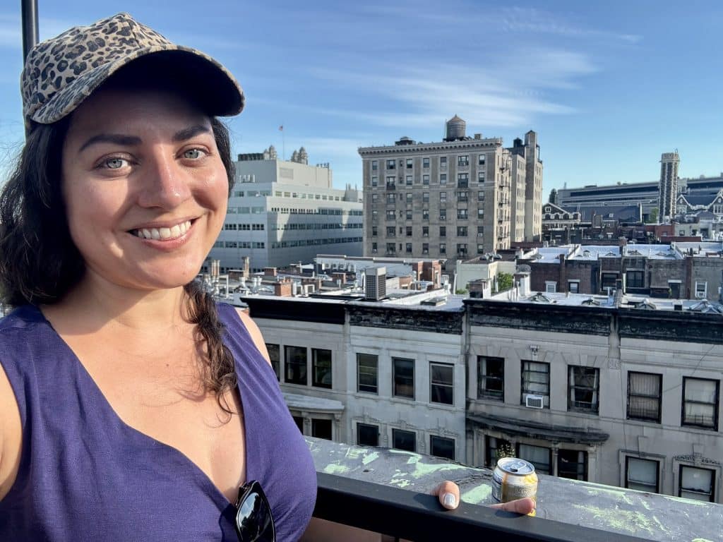 Kate wearing a purple v-neck dress and leopard print baseball cap, and standing on a rooftop in Harlem with city buildings behind her.
