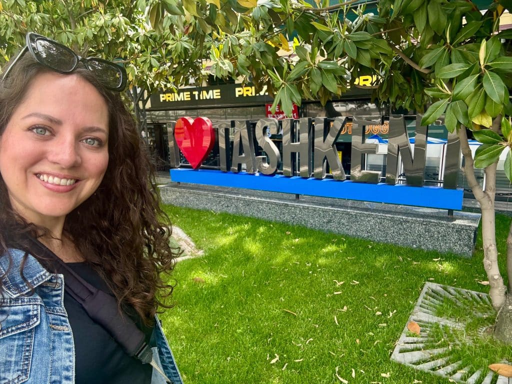 Kate taking a smiling selfie in front of a big sign reading "I Heart Tashkent."