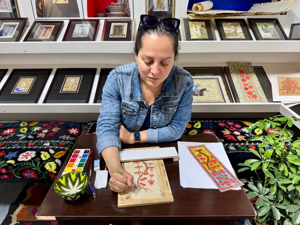 Kate sitting at a table and painting a pomegranate tree on silk paper with watercolors.