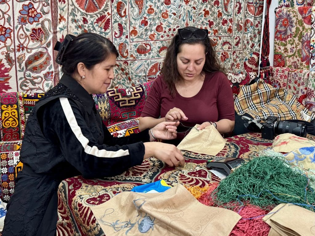 Kate sitting next to an Uzbek woman in her 50s as they embroider on pieces of fabric together.