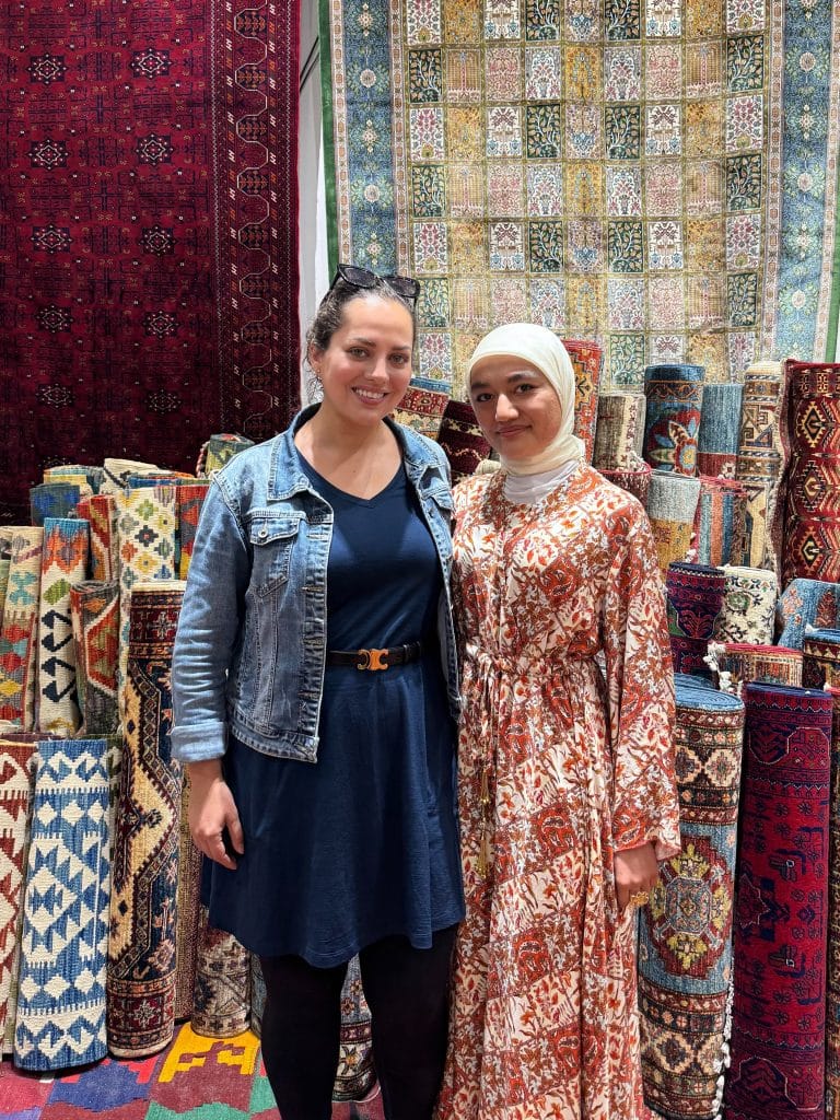 Kate wearing a denim jacket and knee-length dress over leggings with her local friend Muxtarama in a hijab and long red and white patterned dress. They are standing in front of a display of Uzbek rugs.