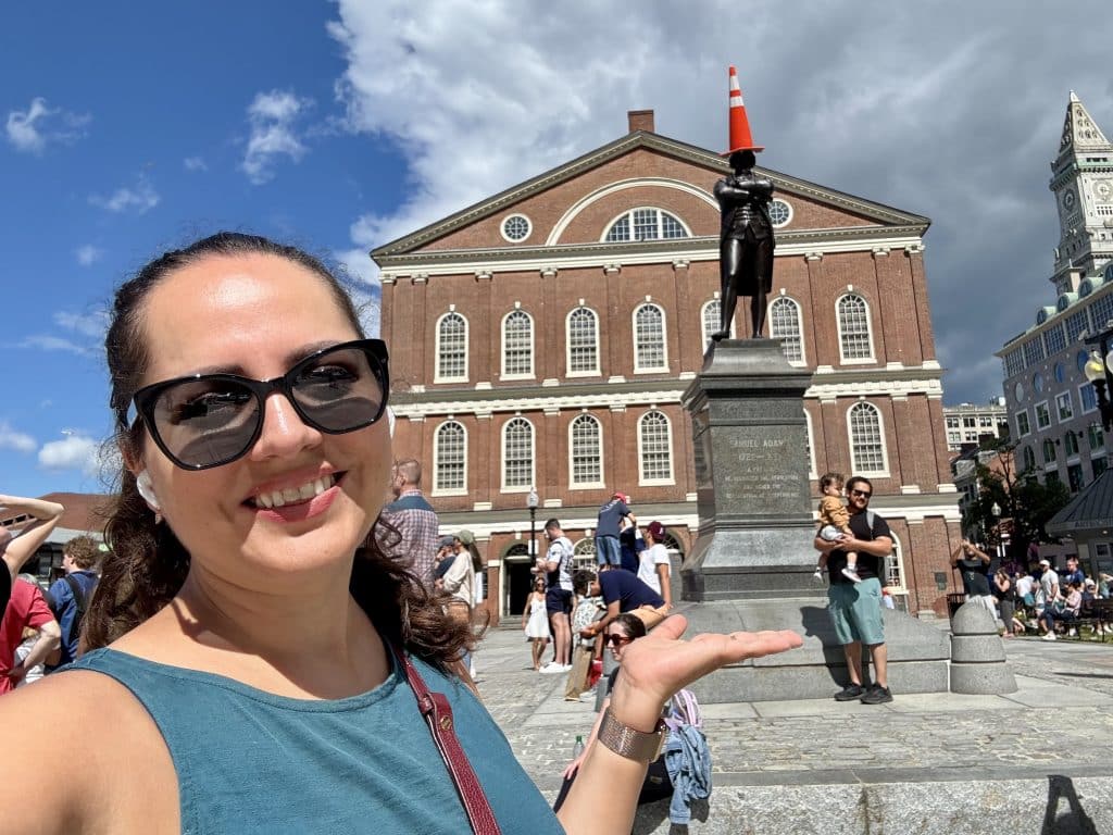 Kate standing in front of the Sam Adams Statue at Faneuil Hall in Boston, with a big orange traffic cone on his head.