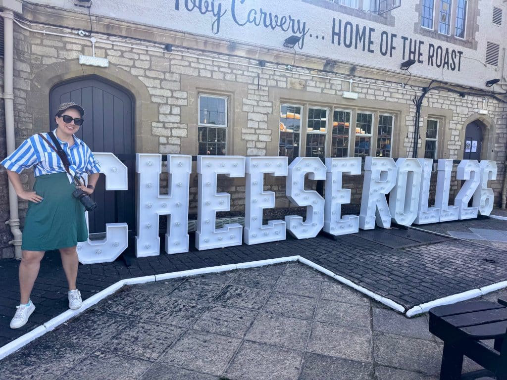 Kate standing in front of a sign reading cheese Roll 26. She wears a dress with a button-down line shirt on top, a baseball cap, sneakers, and sunglasses.