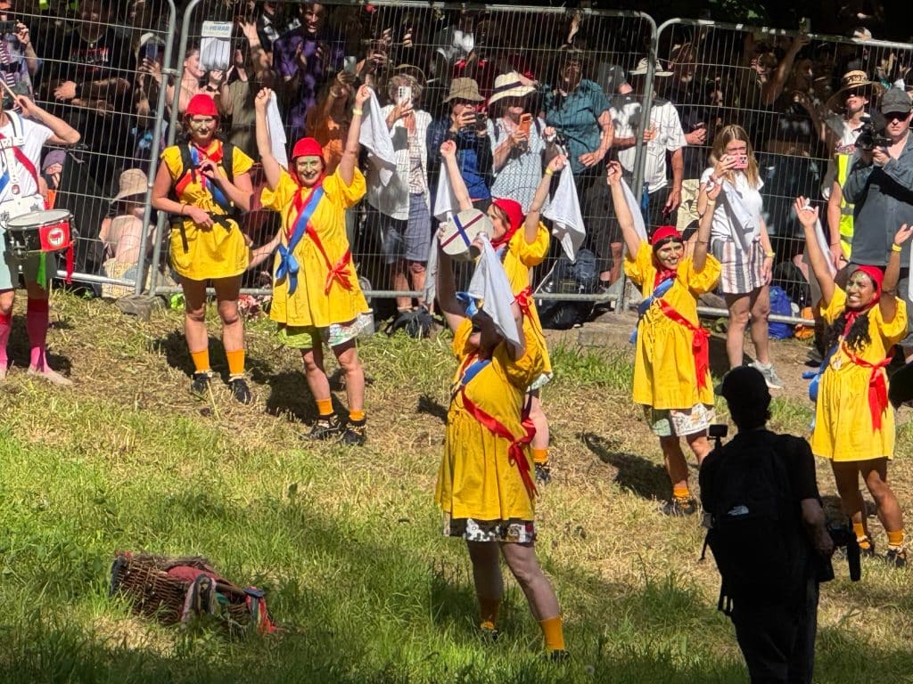 Women in bright yellow and red dresses dancing with handkerchiefs, one holding up a wheel of cheese.