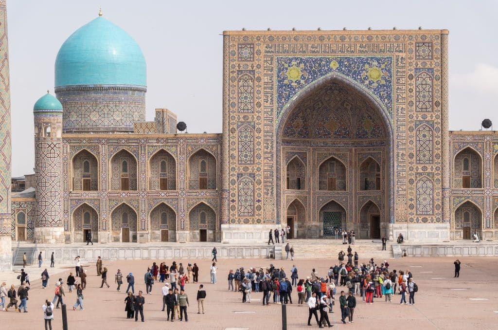 A madrassa with intricately tiled facades and turquoise domes, with lots of people on the square in front of it.