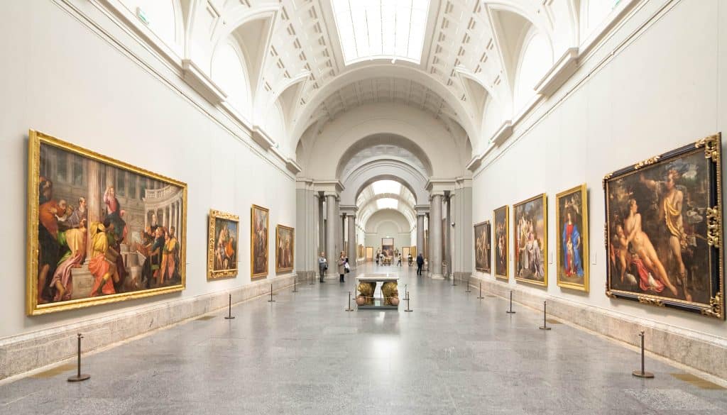 Long perspective of a majestic hallway inside the Prado Museum filled with classical paintings.