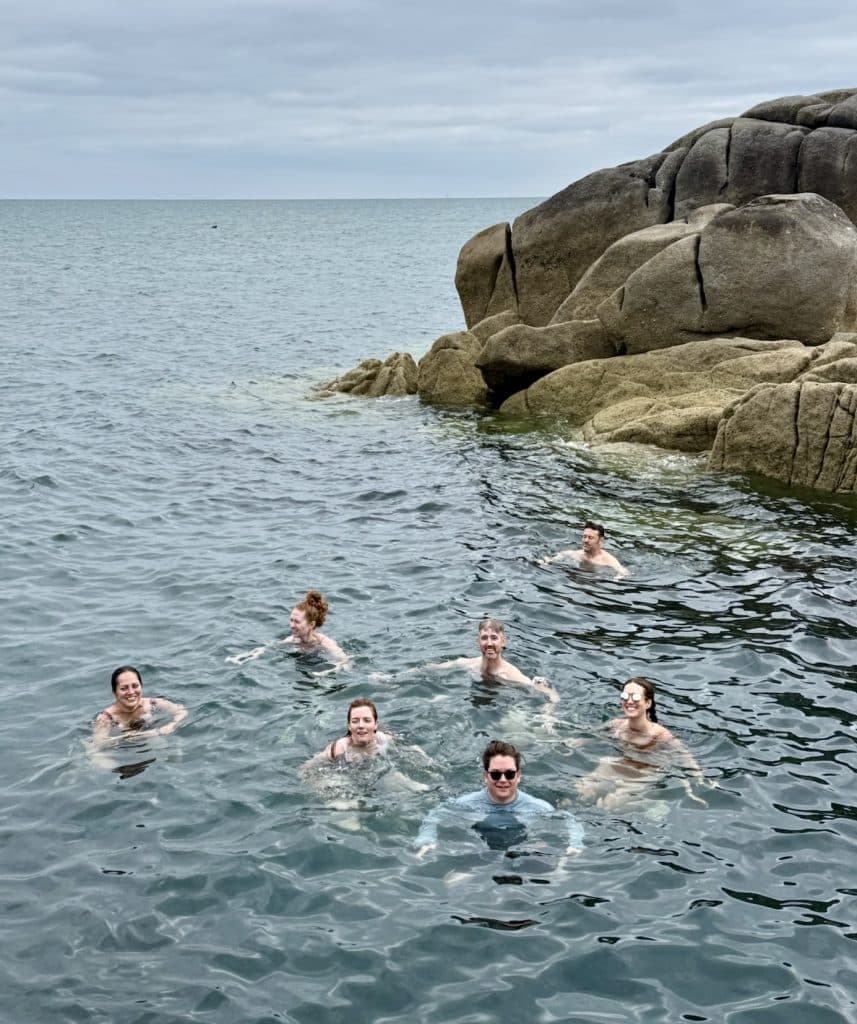 A group of several friends swimming in a rocky area of the Irish Sea, underneath gray clouds.